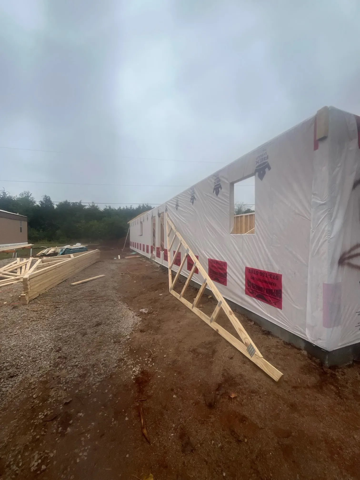 Long house wrapped in weather barrier with a roof truss leaning nearby